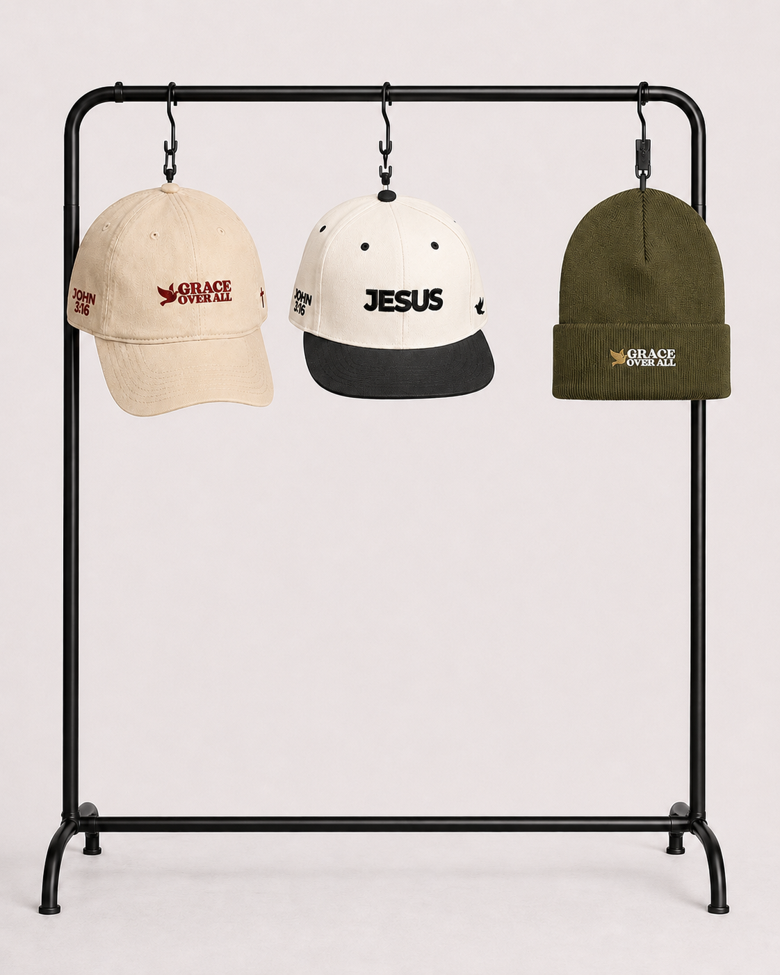 Three caps hanging from a black clothing rack against a light pink background: a beige cap with red 'GRACE SYSTEM' text, a white and black cap with 'JESUS' in black, and an olive green beanie with a small 'GRACE SYSTEM' logo.