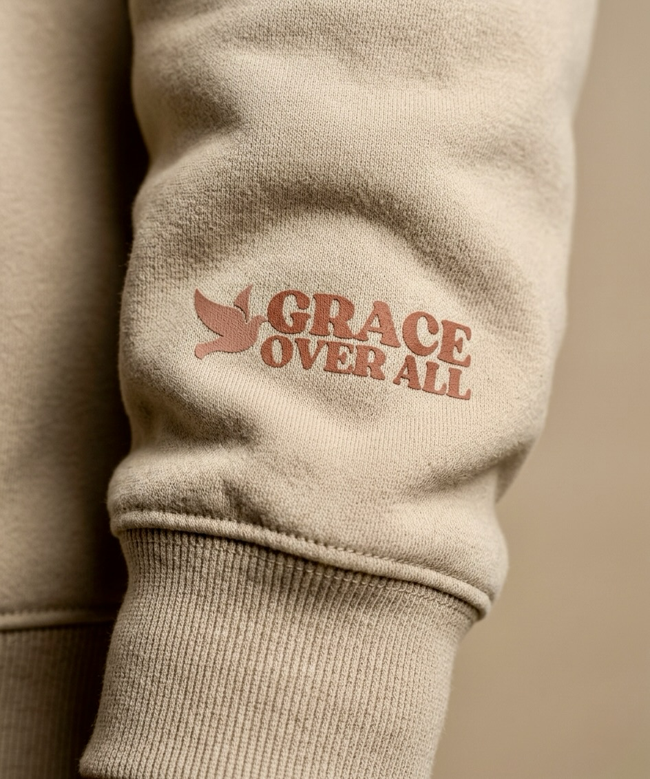 Close-up of Grace Over All beige hoodie sleeve featuring dove logo and text detail, highlighting premium fabric and faith-based design.
